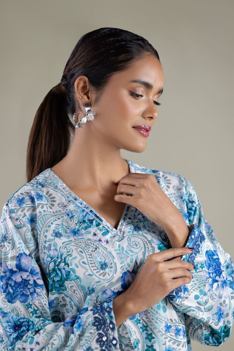 A model wearing a 1-26-143-A-E 2-piece lawn suit by Khaadi. The outfit features a long kurta with intricate vertical patterns and detailed borders, paired with dyed trousers and a matching printed dupatta with striped and floral designs. The model stands against a plain background, holding the dupatta, with product fabric details displayed on the left side.