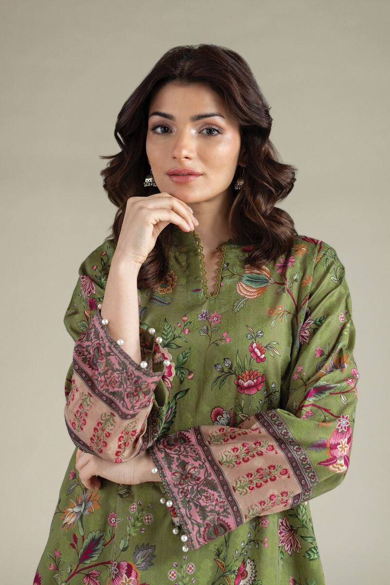 A model wearing a A112-26-101ED2 2-piece lawn suit by Khaadi. The outfit features a long kurta with intricate vertical patterns and detailed borders, paired with dyed trousers and a matching printed dupatta with striped and floral designs. The model stands against a plain background, holding the dupatta, with product fabric details displayed on the left side.
