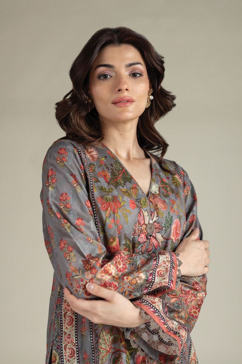 A model wearing a A112-26-101EE2 2-piece lawn suit by Khaadi. The outfit features a long kurta with intricate vertical patterns and detailed borders, paired with dyed trousers and a matching printed dupatta with striped and floral designs. The model stands against a plain background, holding the dupatta, with product fabric details displayed on the left side.