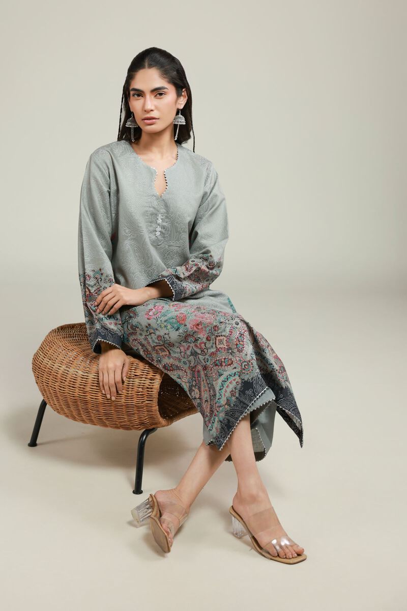 A woman wearing a grey printed kurta with floral patterns near the hem and sleeves, paired with matching straight trousers and transparent heels.